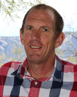 A man wearing a red and blue plaid shirt smiles at the camera. The background shows blurred greenery and a faint view of hills or mountains under a partly cloudy sky.